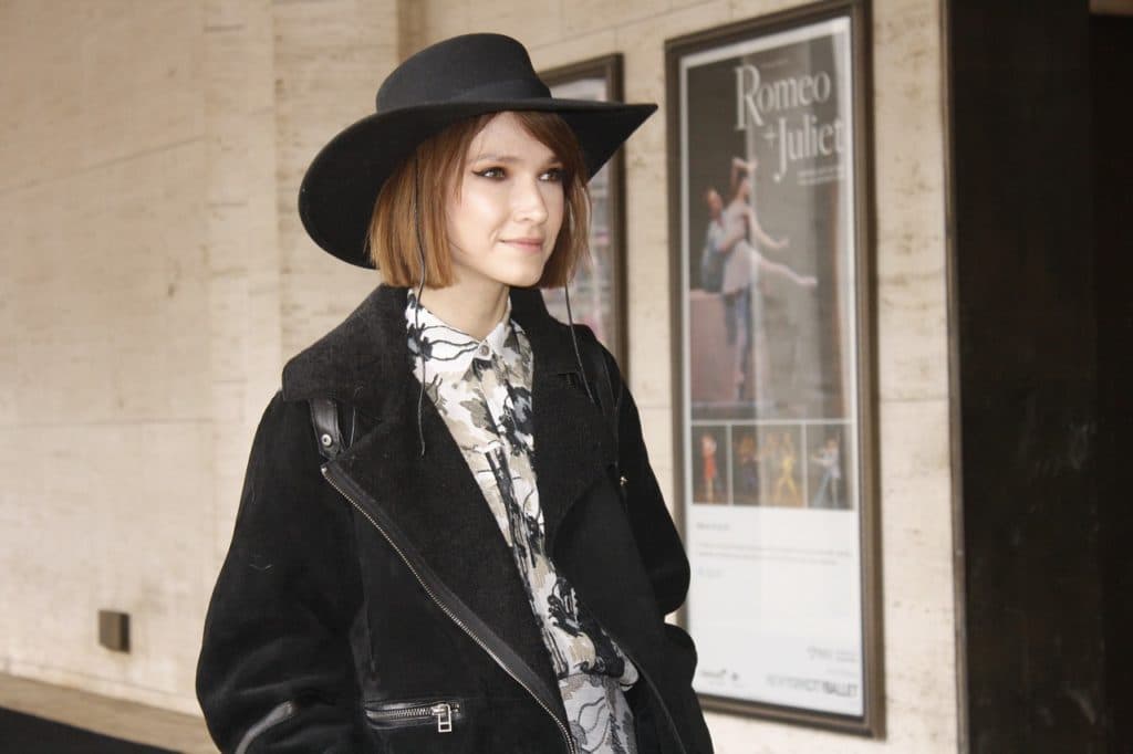 A woman in a black wide brimmed hat and shearling jacket beside a theatre poster