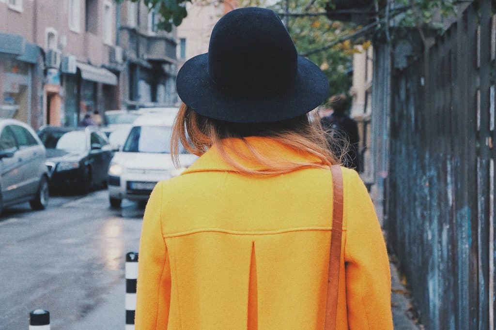 A woman in a mustard yellow coat and black hat walking away down a city street