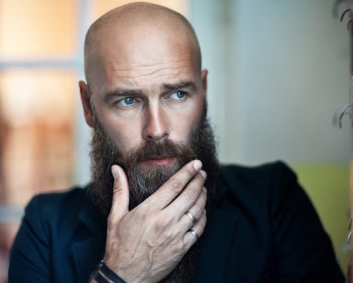 Portrait of a bald man with a full beard resting a hand on his chin