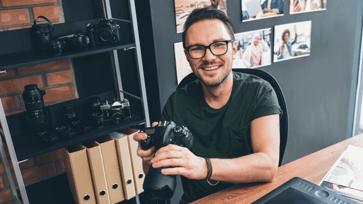 A photographer holding a camera at a desk lined with lenses and equipment